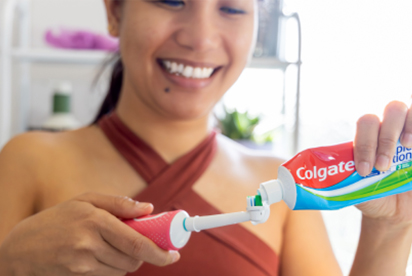 Mujer sonriente aplicando pasta de dientes Colgate en un cepillo eléctrico rosa para su rutina de higiene bucal.